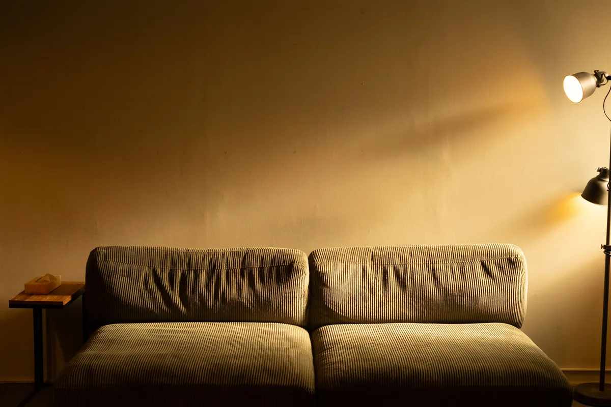 An empty corduroy sofa in a dim living room, lit by a single floor lamp casting a warm glow up the wall.